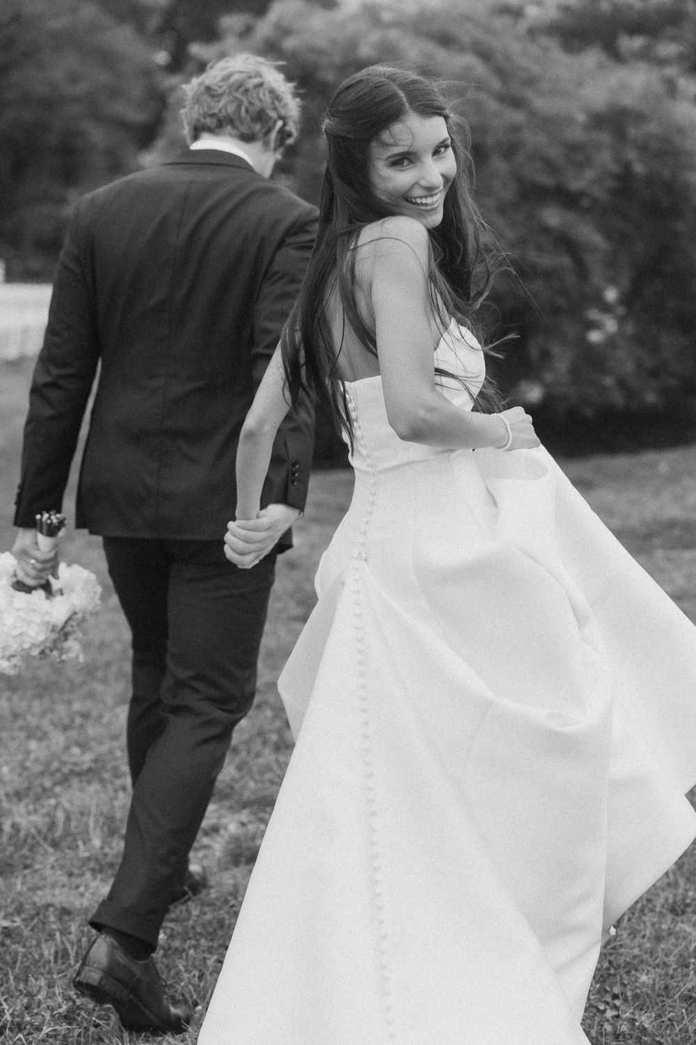 Black and white photo of smiling bride in white gown looking back while holding grooms hand outdoors.