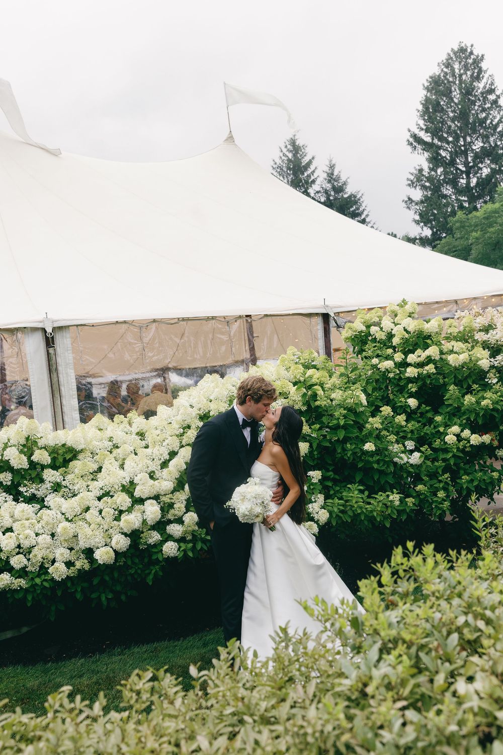 Bride and groom share a kiss surrounded by white hydrangeas beside an elegant wedding tent outdoors.