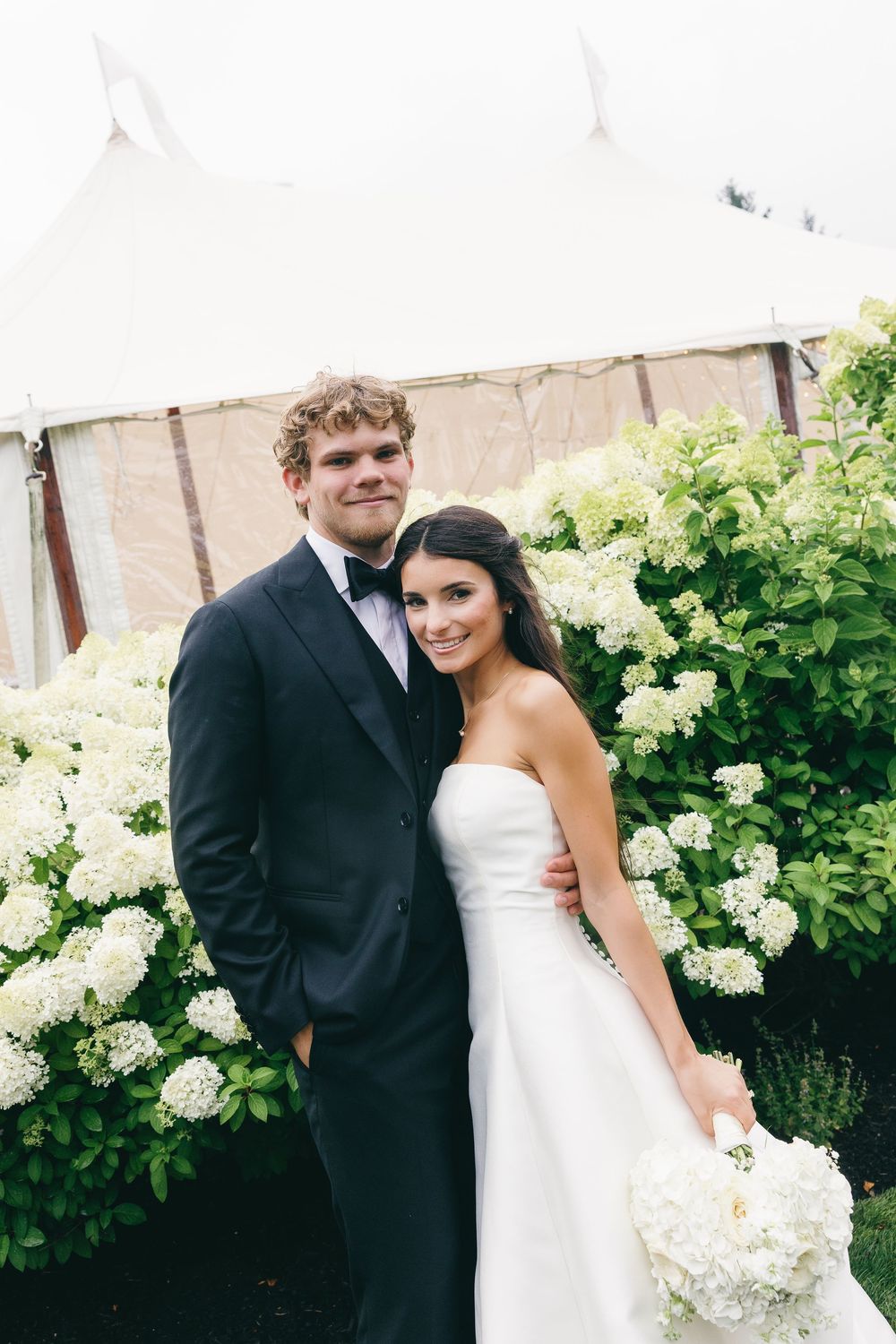 Bride and groom posing in front of white hydrangea bushes and a wedding tent, bride holding a bouquet.