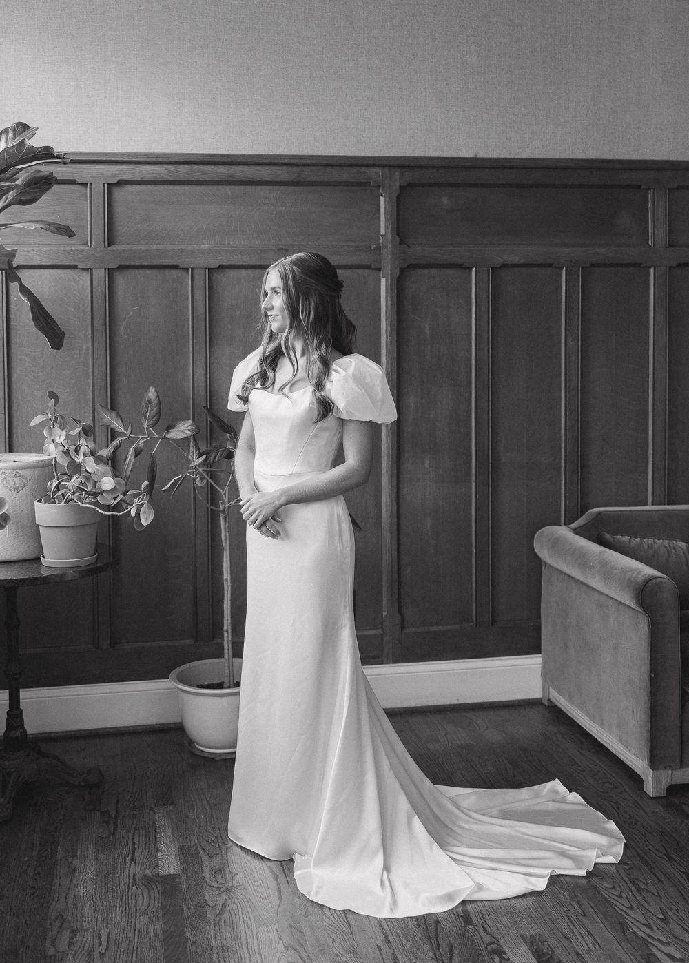 Black and white photo of a bride in a flowing wedding gown with puff sleeves standing in an elegant wood-paneled room.
