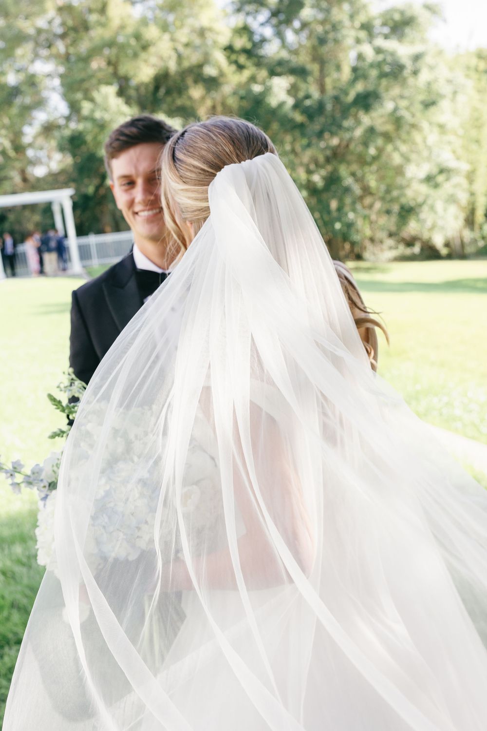 Smiling groom in black tuxedo lifting bride's flowing white veil outdoors in a green garden setting.