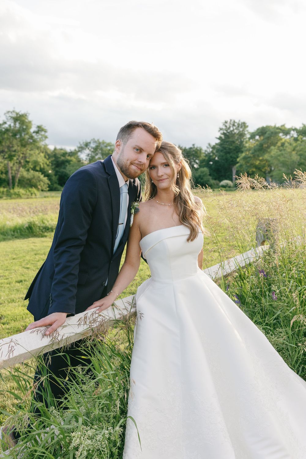Bride and groom pose by a rustic wooden fence in a lush green field on their wedding day.