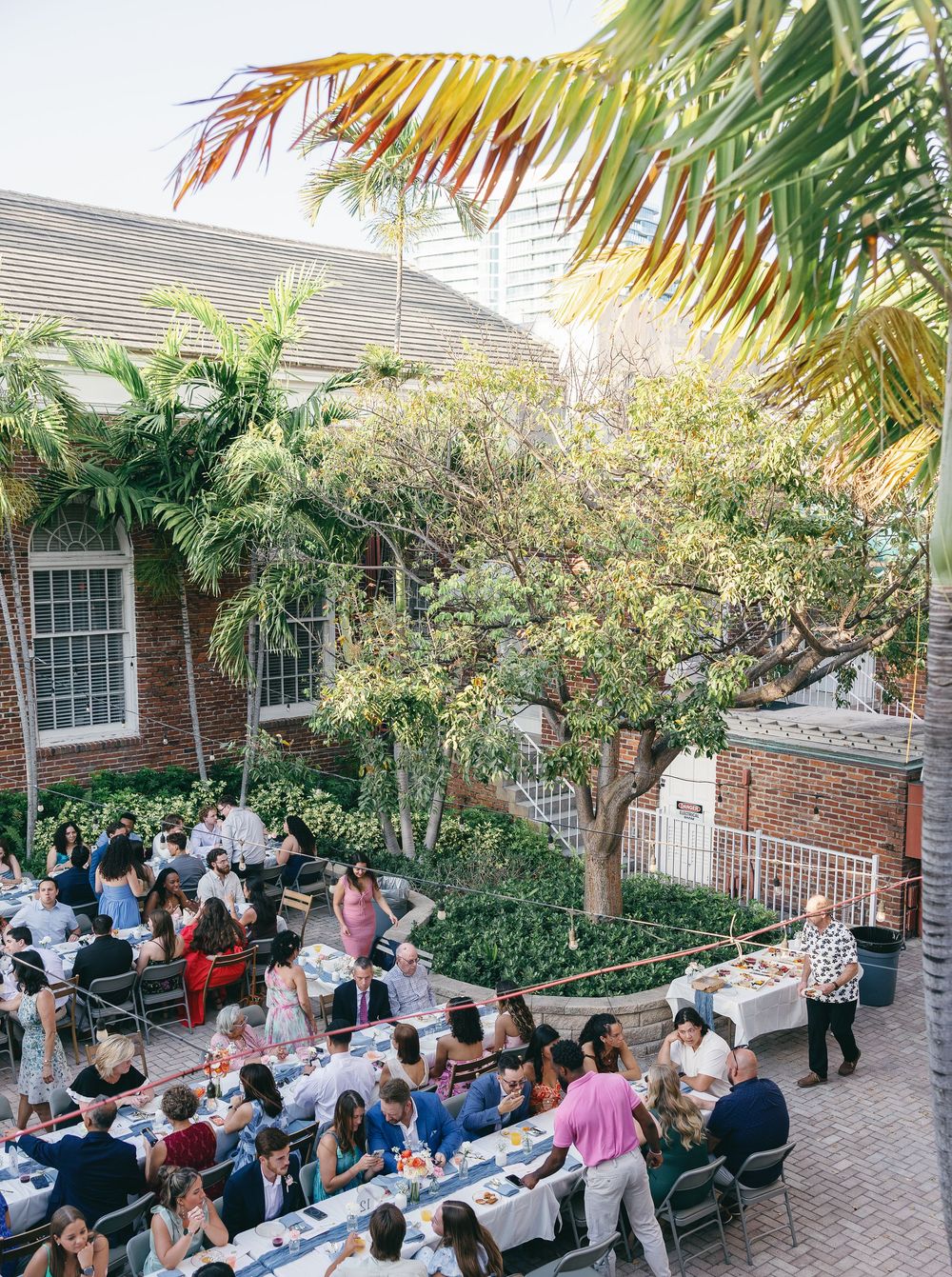 Guests dining at long tables in a tropical courtyard beside a brick building with palm trees overhead.
