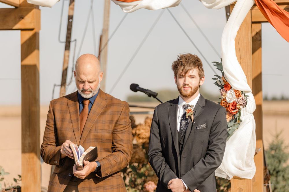 An outdoor wedding ceremony takes place under a wooden pergola with warm autumn sunlight and rustic decor.