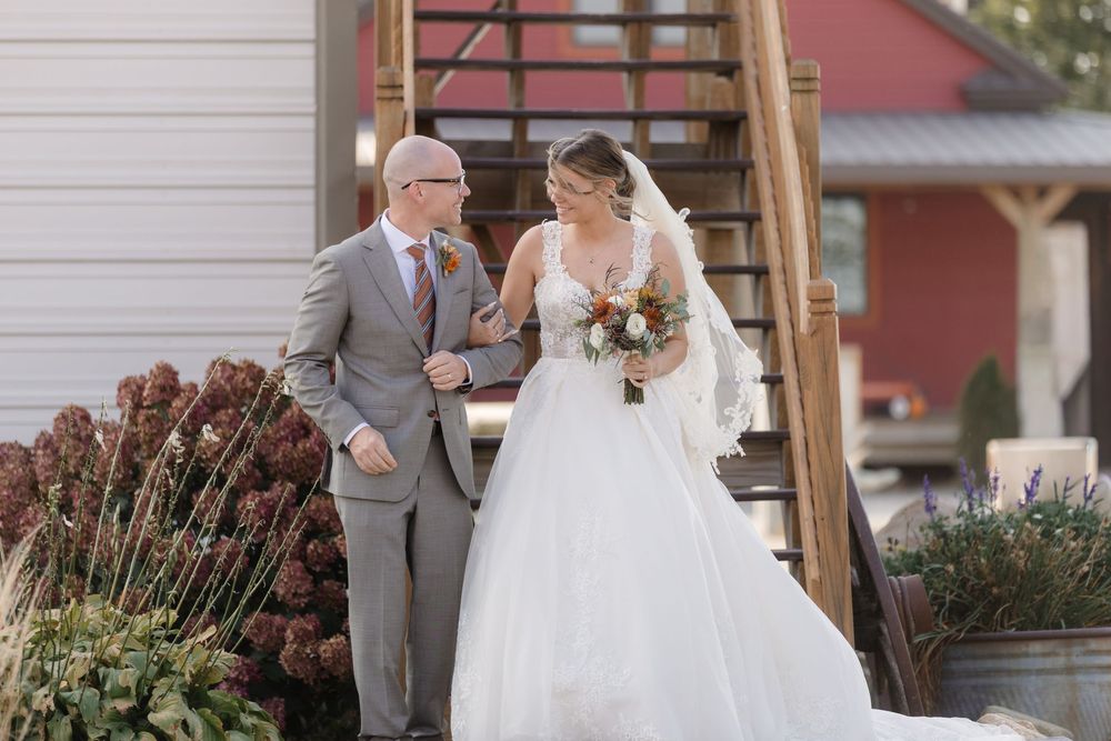 A bride in a white wedding gown descends outdoor stairs with a companion in a grey suit on a sunny day.