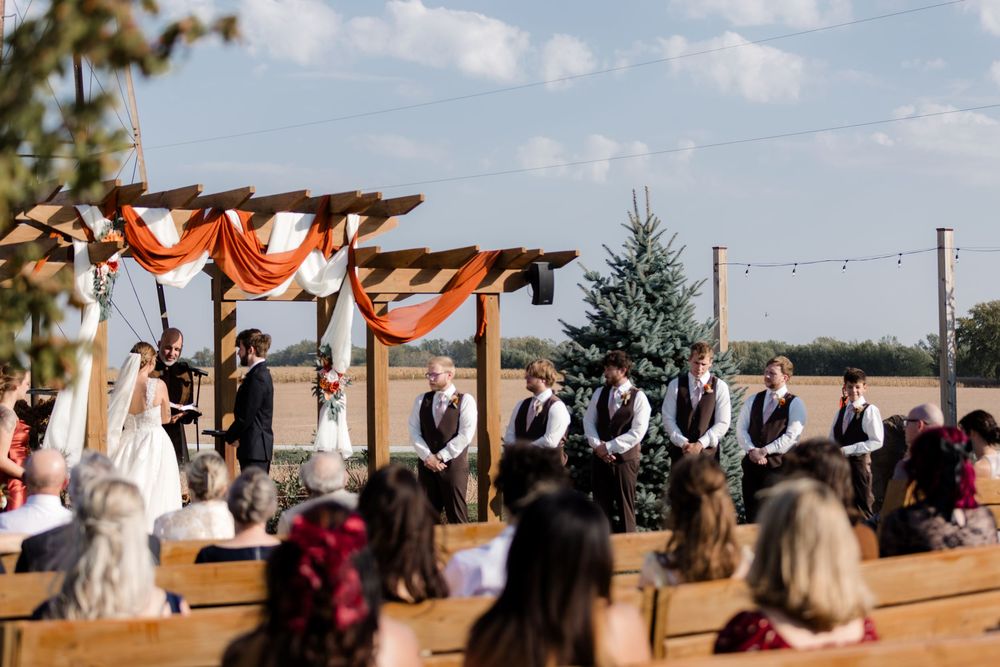 Outdoor wedding ceremony setup with orange and white fabric draped over wooden pergola and white chairs on grass.