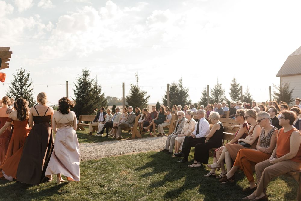 Wedding guests seated on chairs outdoors during a sunset ceremony with trees in the background.