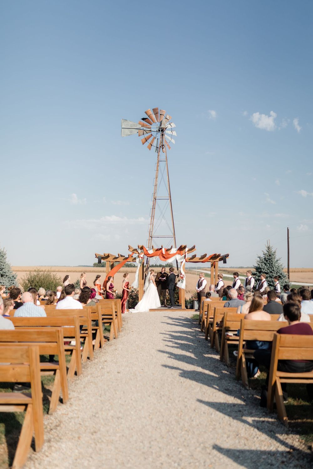Outdoor wedding ceremony setup with wooden benches and a rustic windmill backdrop under blue skies.