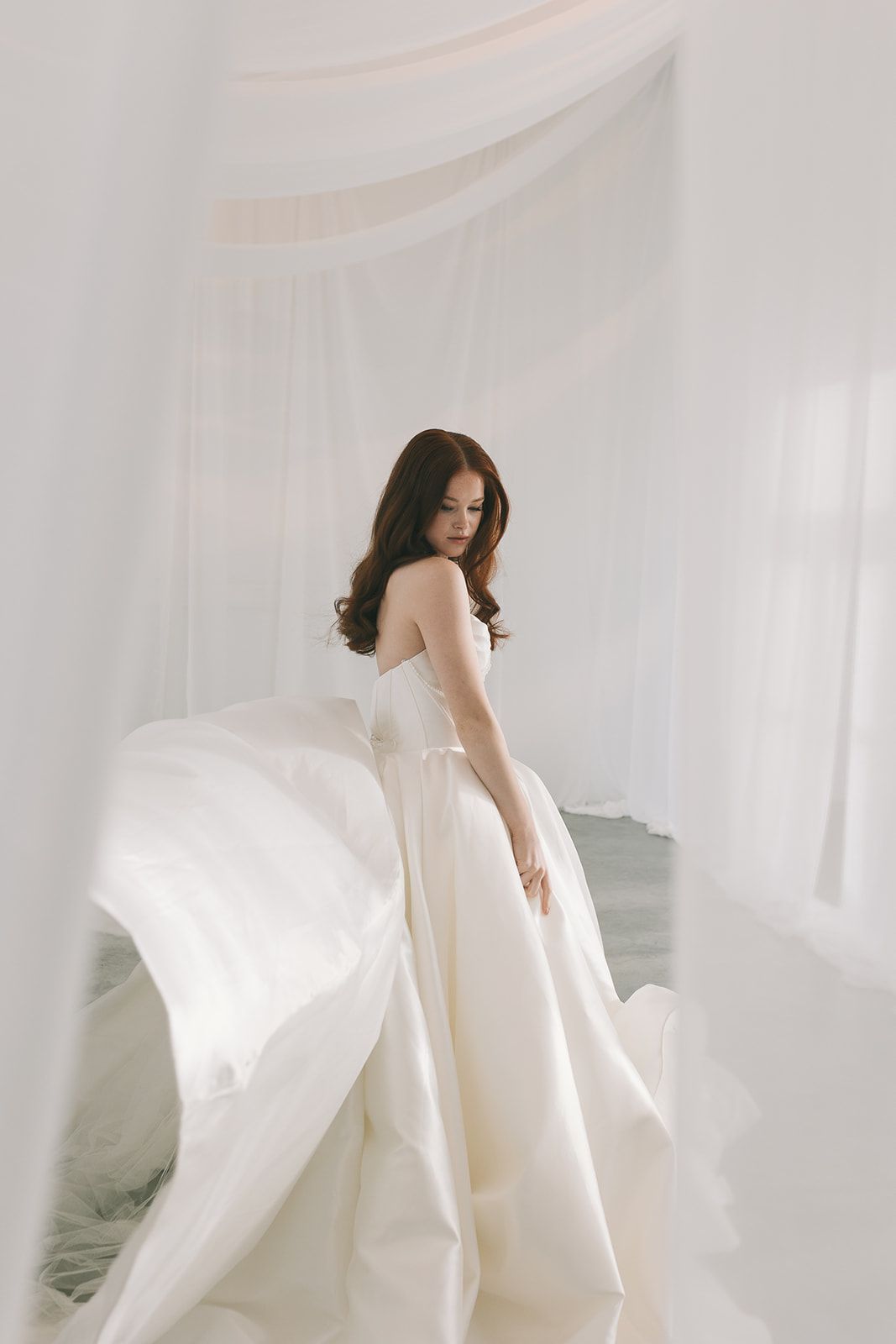 Ethereal bridal photoshoot featuring flowing white veil and dress against soft gray background.