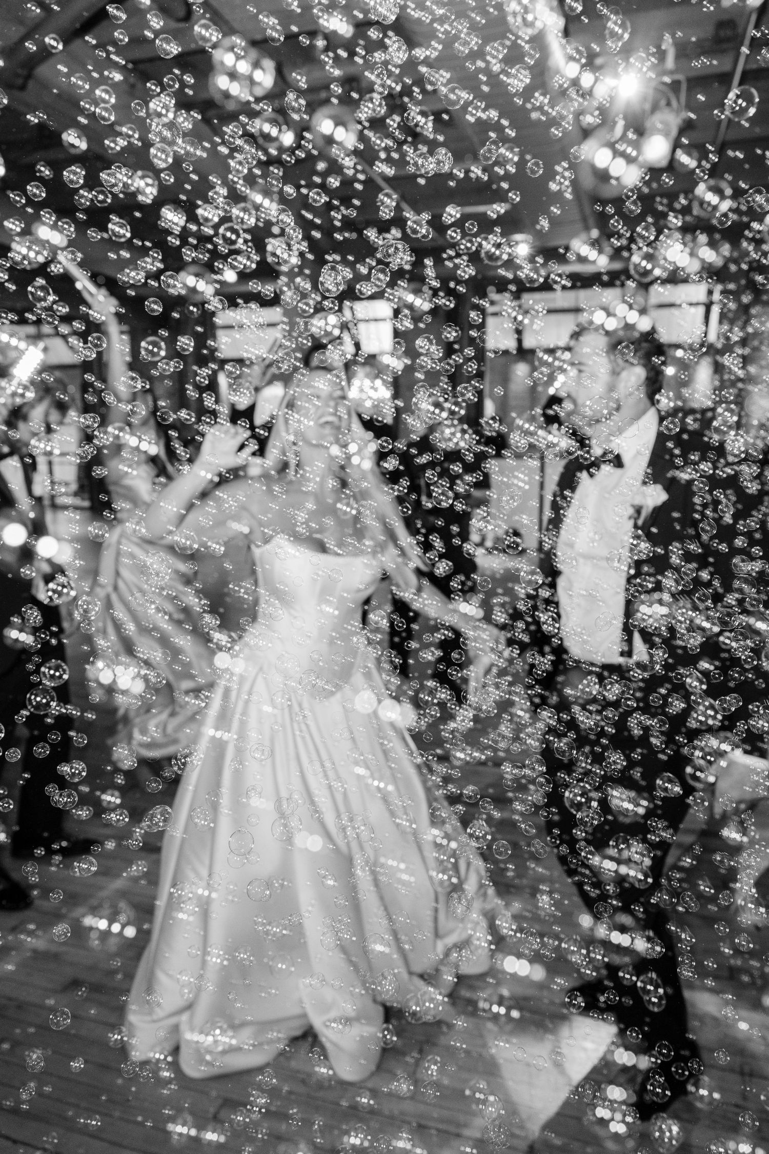 A couple dances joyfully surrounded by floating soap bubbles on a wooden dance floor at an indoor celebration.