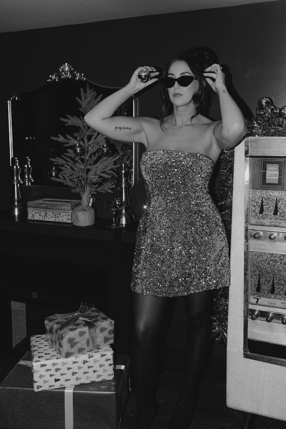 A person in a sparkly dress poses dramatically in a dark room with holiday decor and wrapped presents.