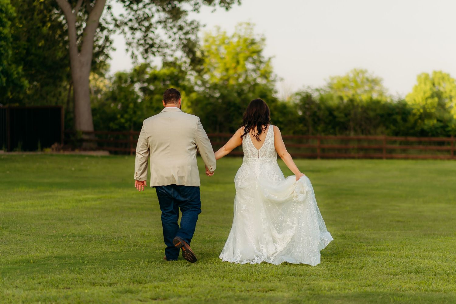 A couple walks hand in hand across a lush green lawn during their outdoor wedding celebration.