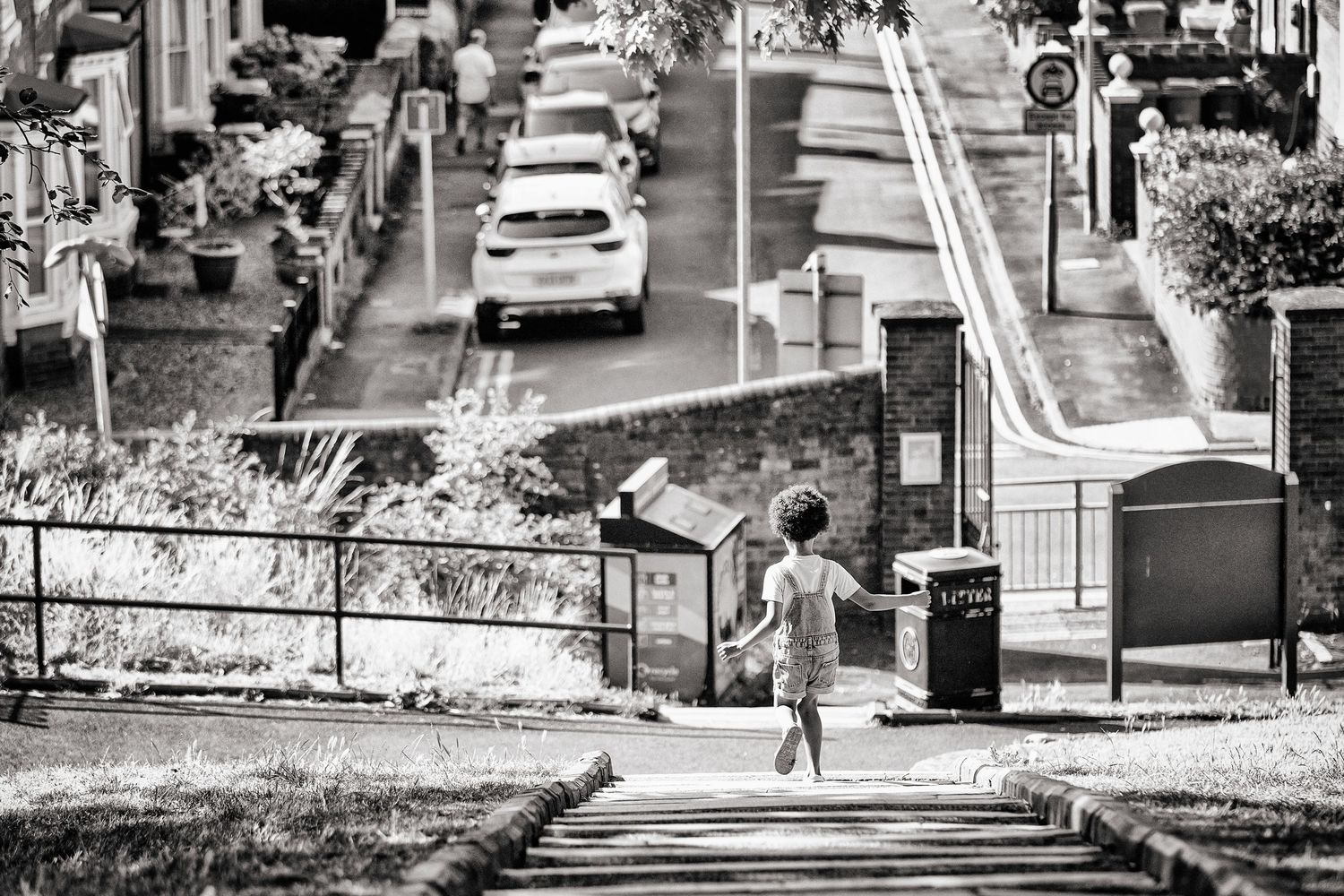 A black and white urban street scene shows a child running down steep stairs in a park.