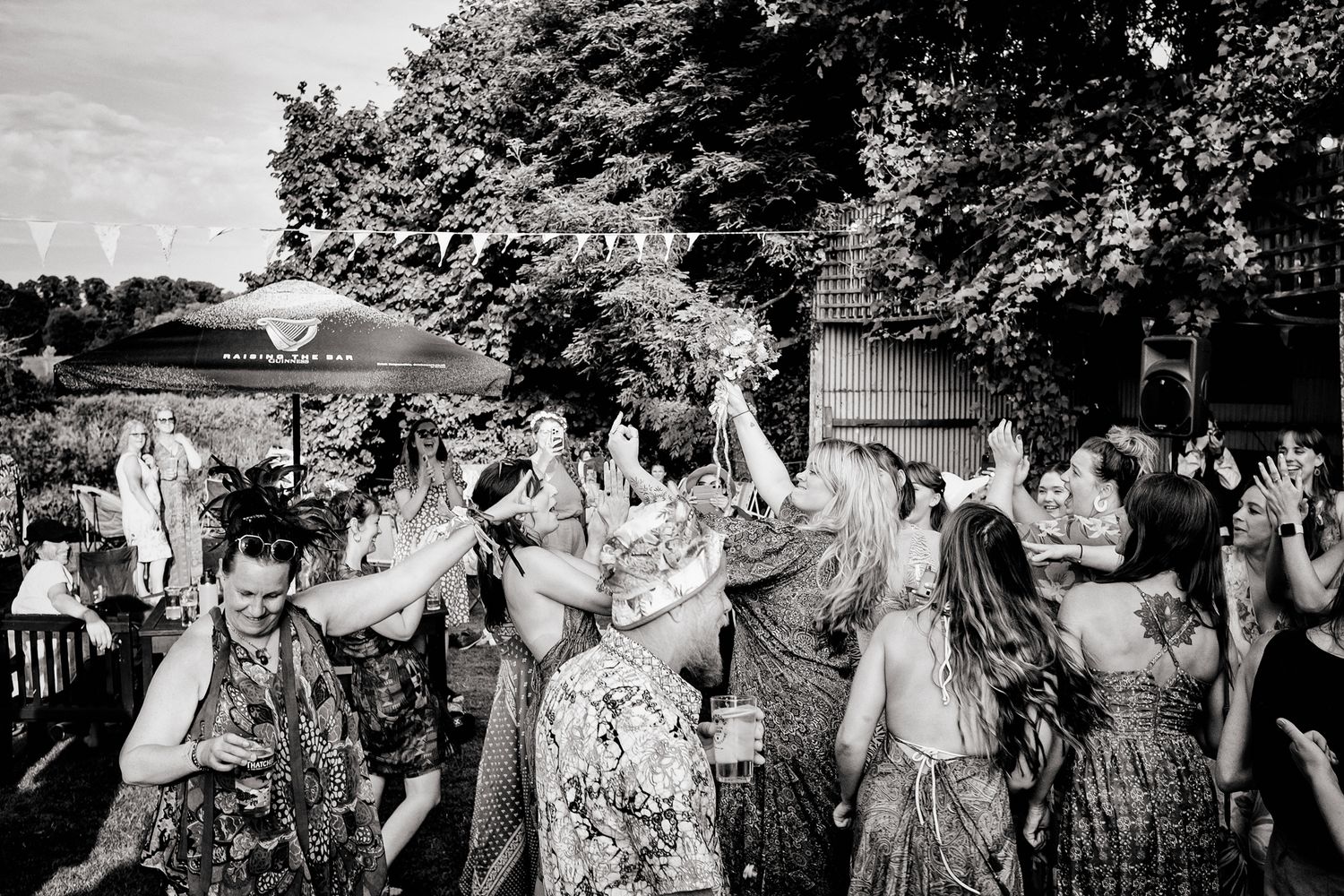 Black and white photograph of guests enjoying an outdoor garden wedding reception with umbrellas and trees in summer.
