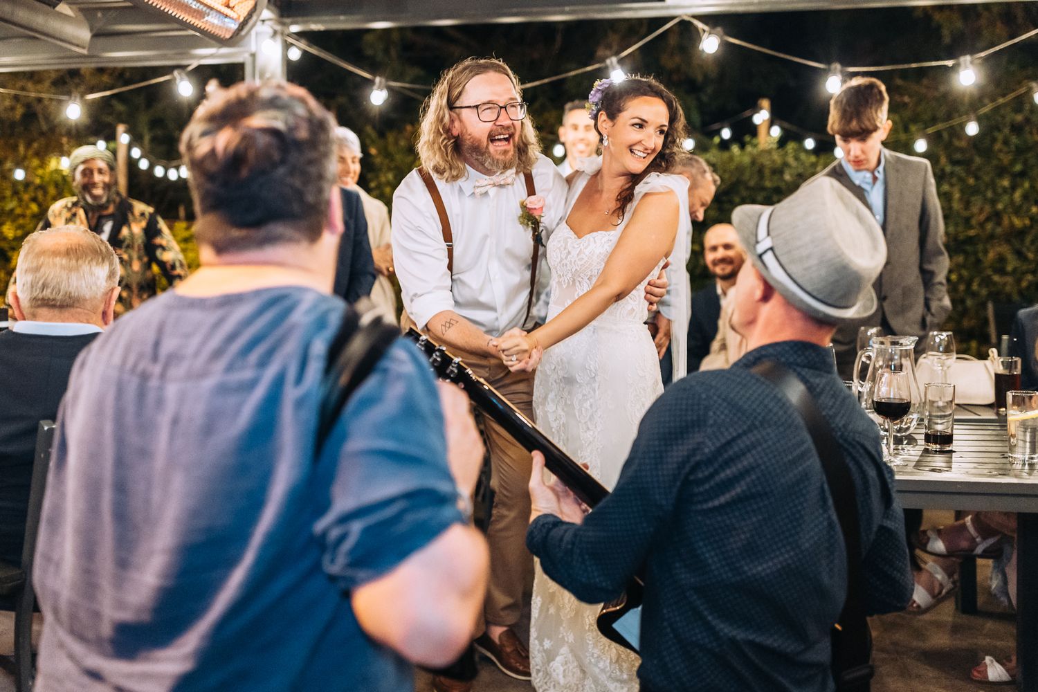 Wedding celebration at an outdoor venue with string lights and guests gathered around tables.