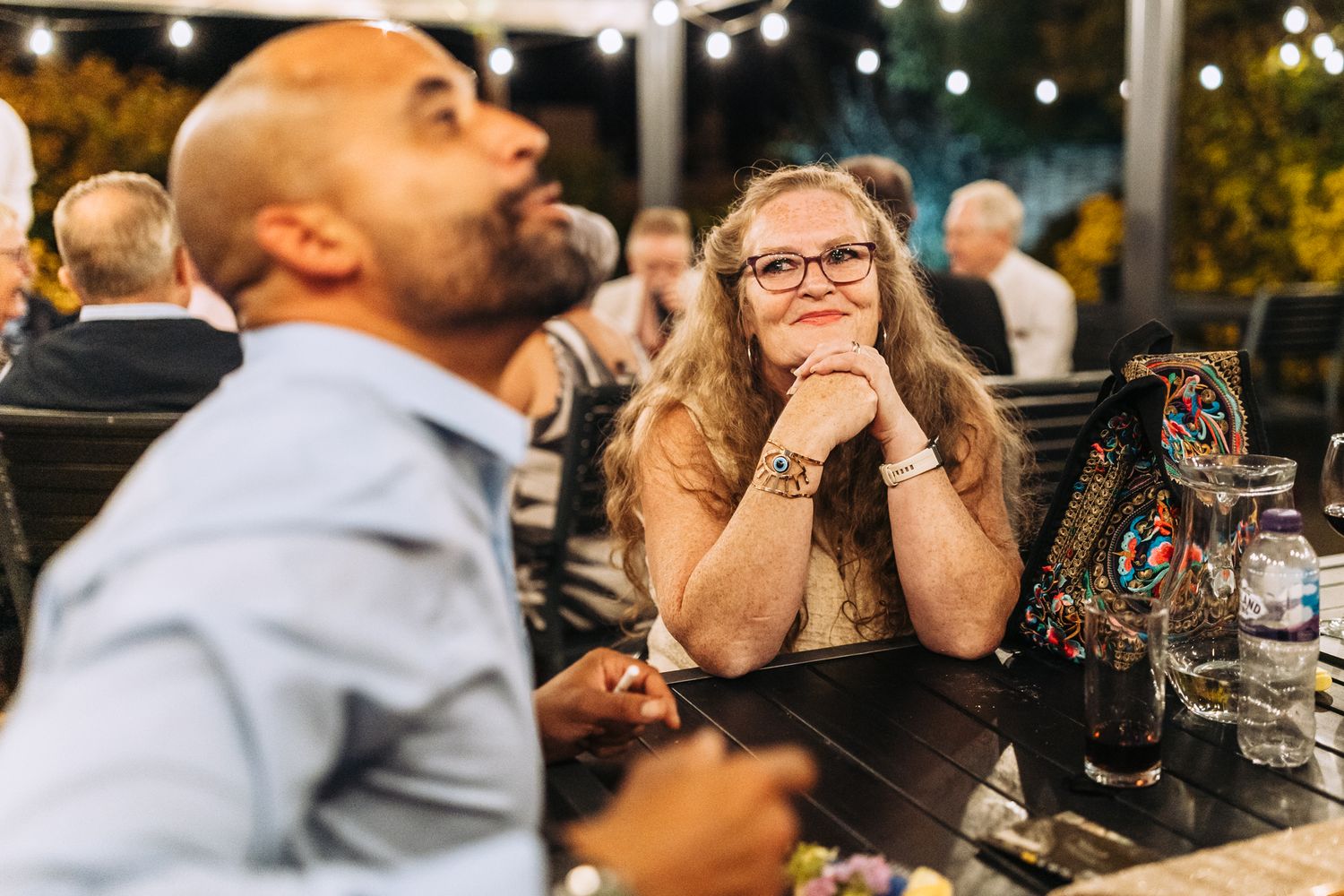 Guests enjoying a lively outdoor evening reception with string lights and drinks at a table.