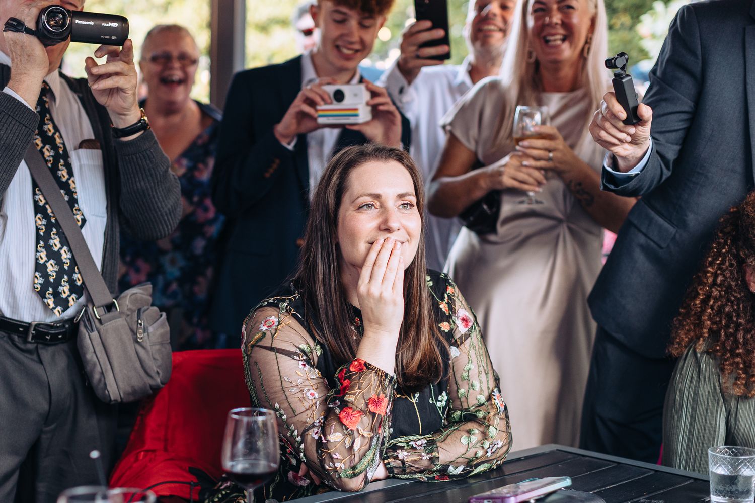 A woman in a floral dress reacts emotionally while surrounded by people holding phones at an event.