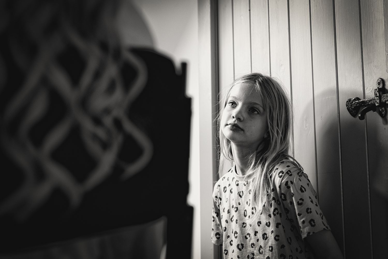 A black and white portrait of a young child with long blonde hair standing near a wall in dramatic lighting.