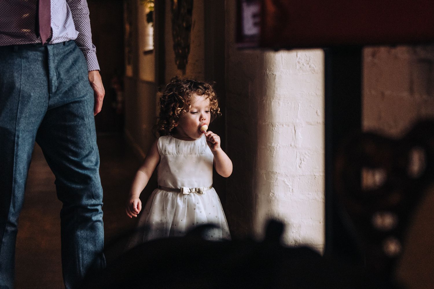 A small child in a white dress sucking a lollypop stands in dramatic lighting casting shadows on a wall.