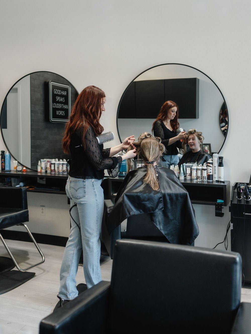 A hairstylist blow-drying a client's hair in a modern salon with round mirrors and professional hair products.
