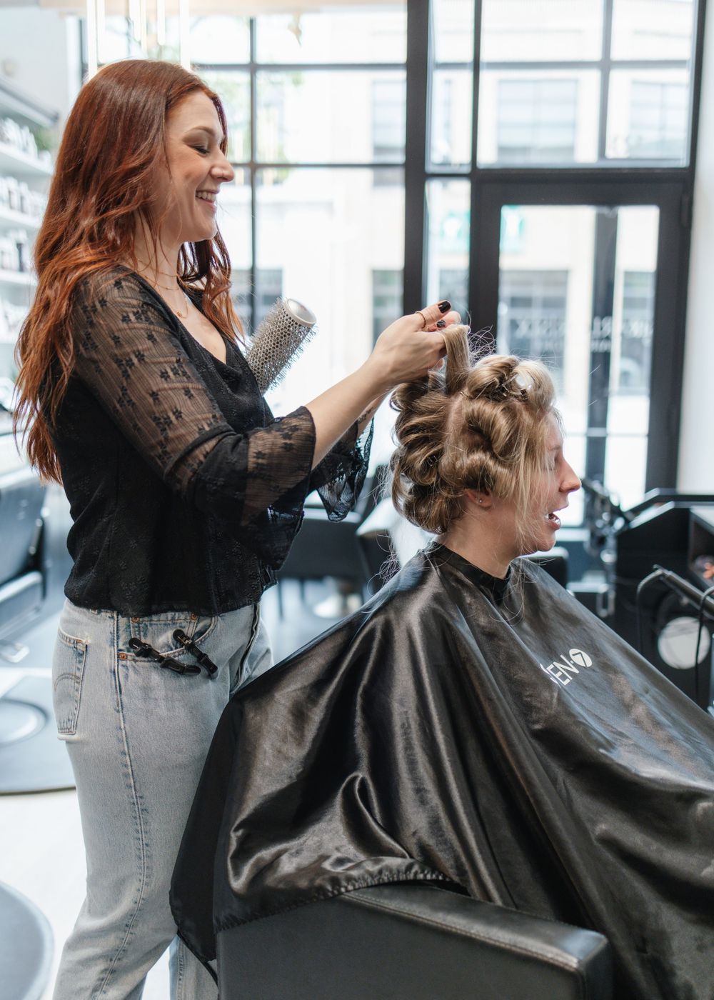 Smiling red-haired stylist styling a client's hair in a modern salon.