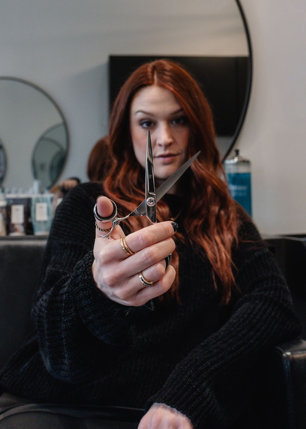 Female hairstylist with long red hair holding scissors toward camera in a professional salon setting.