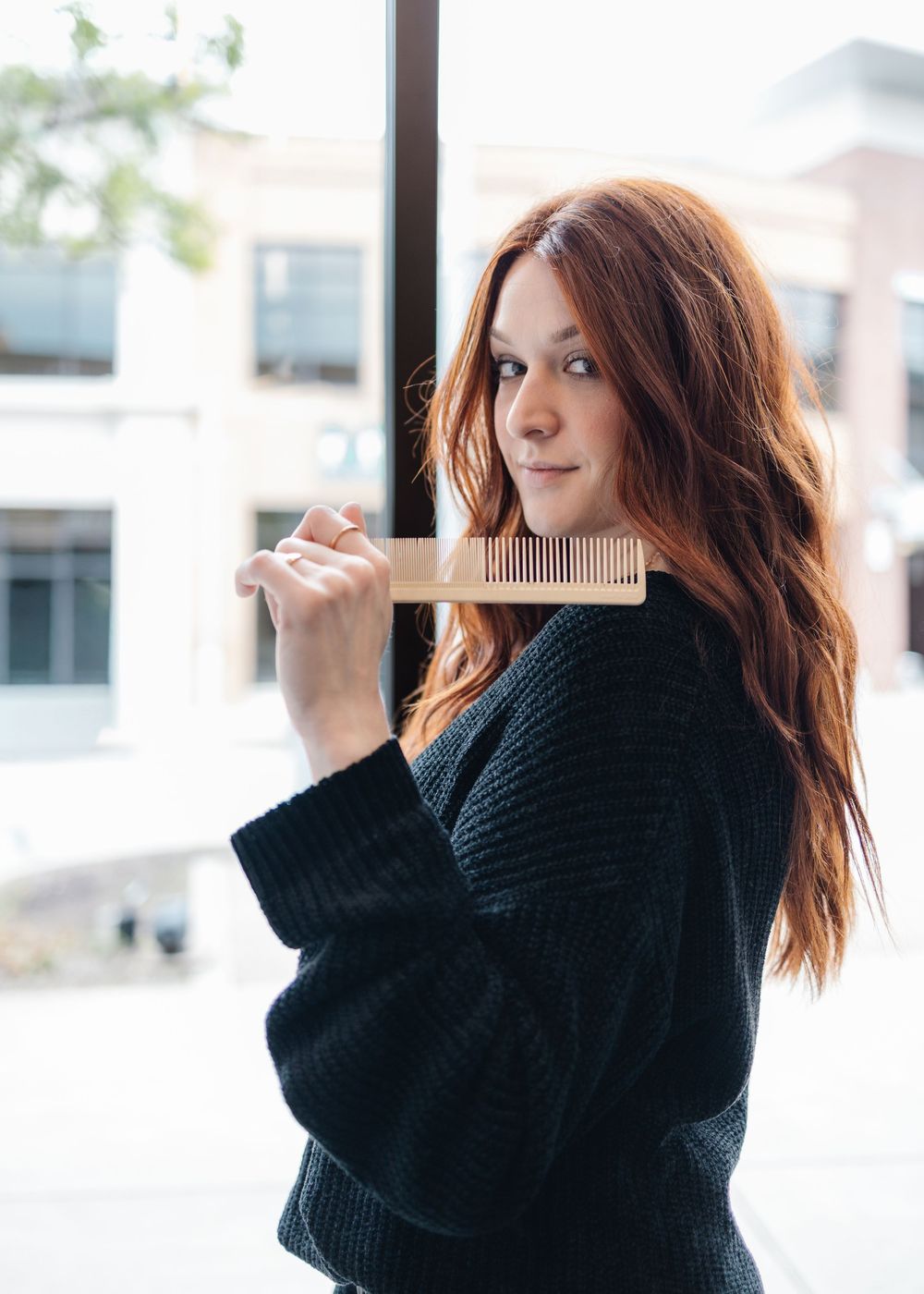 Young woman with long red hair holding a wooden comb in a sweater.