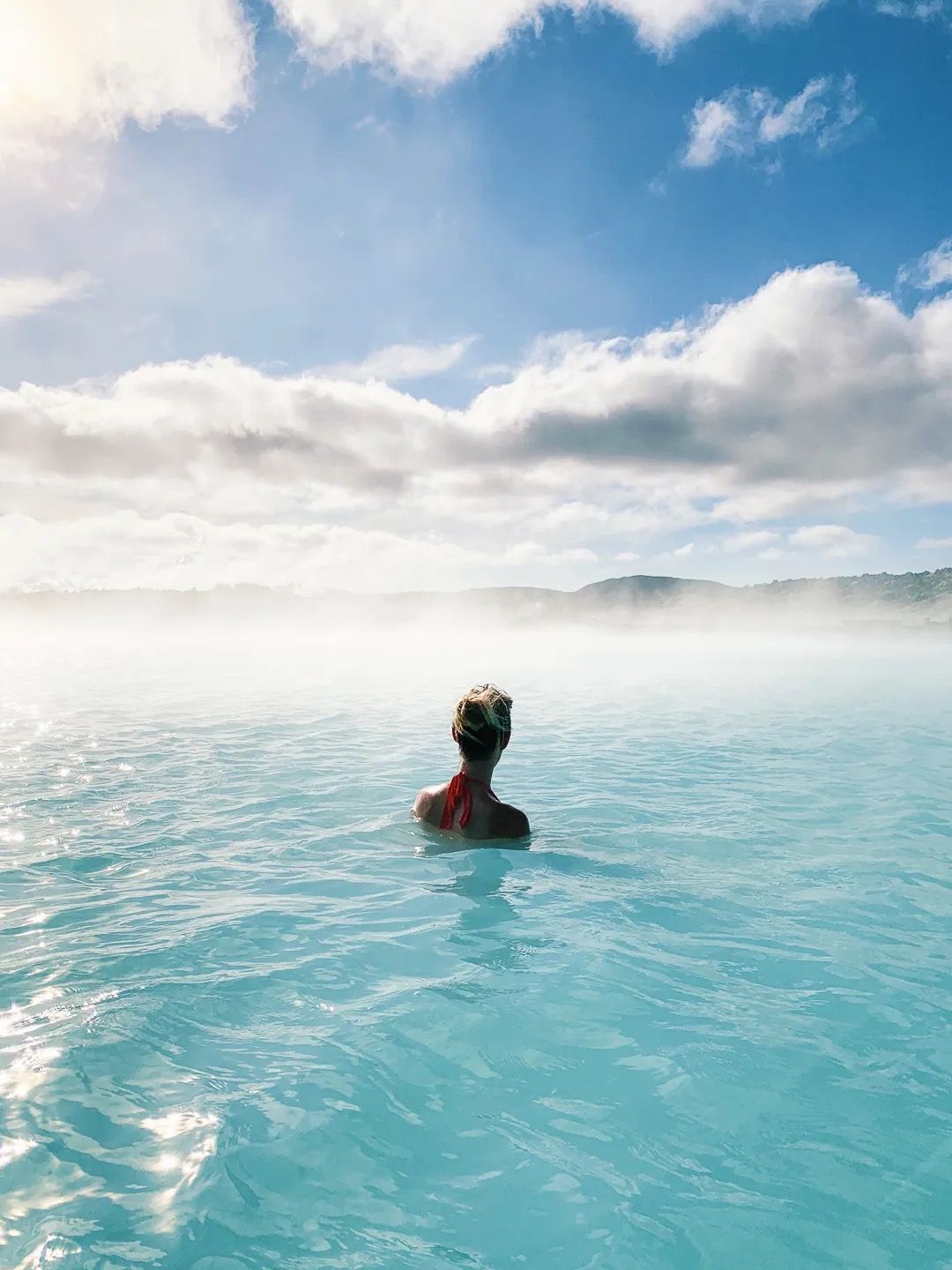 Person relaxing in bright blue geothermal water under a cloudy sky.