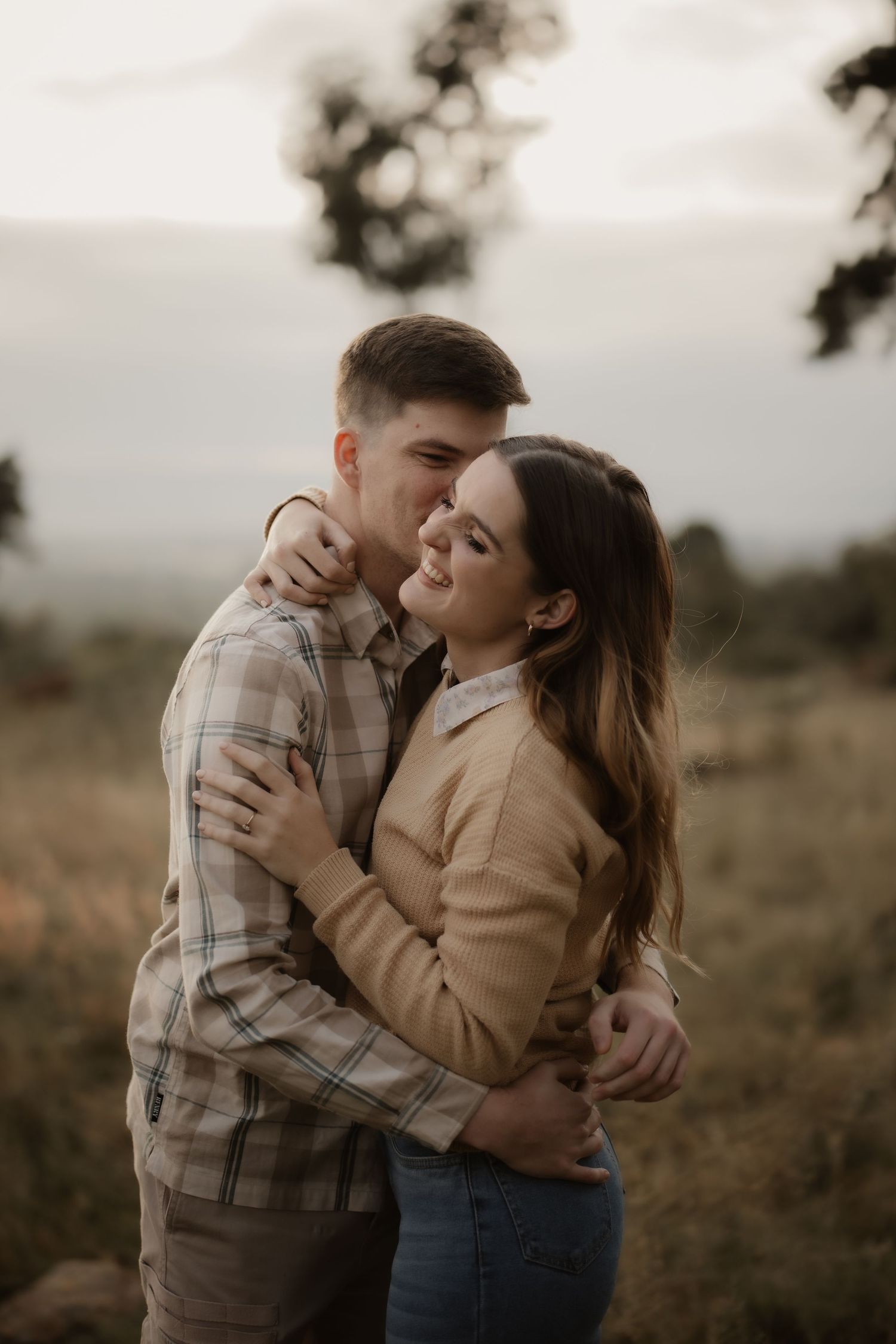 A tender embrace between a couple in casual attire during sunset in a rural setting.