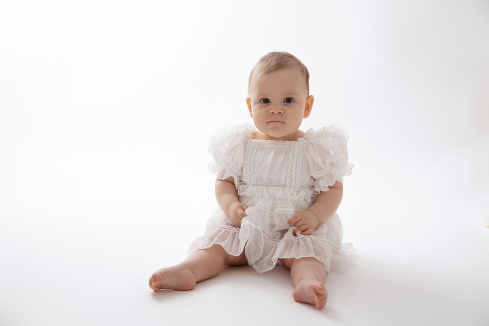 Baby wearing white lace christening gown sits on white background in four different poses.