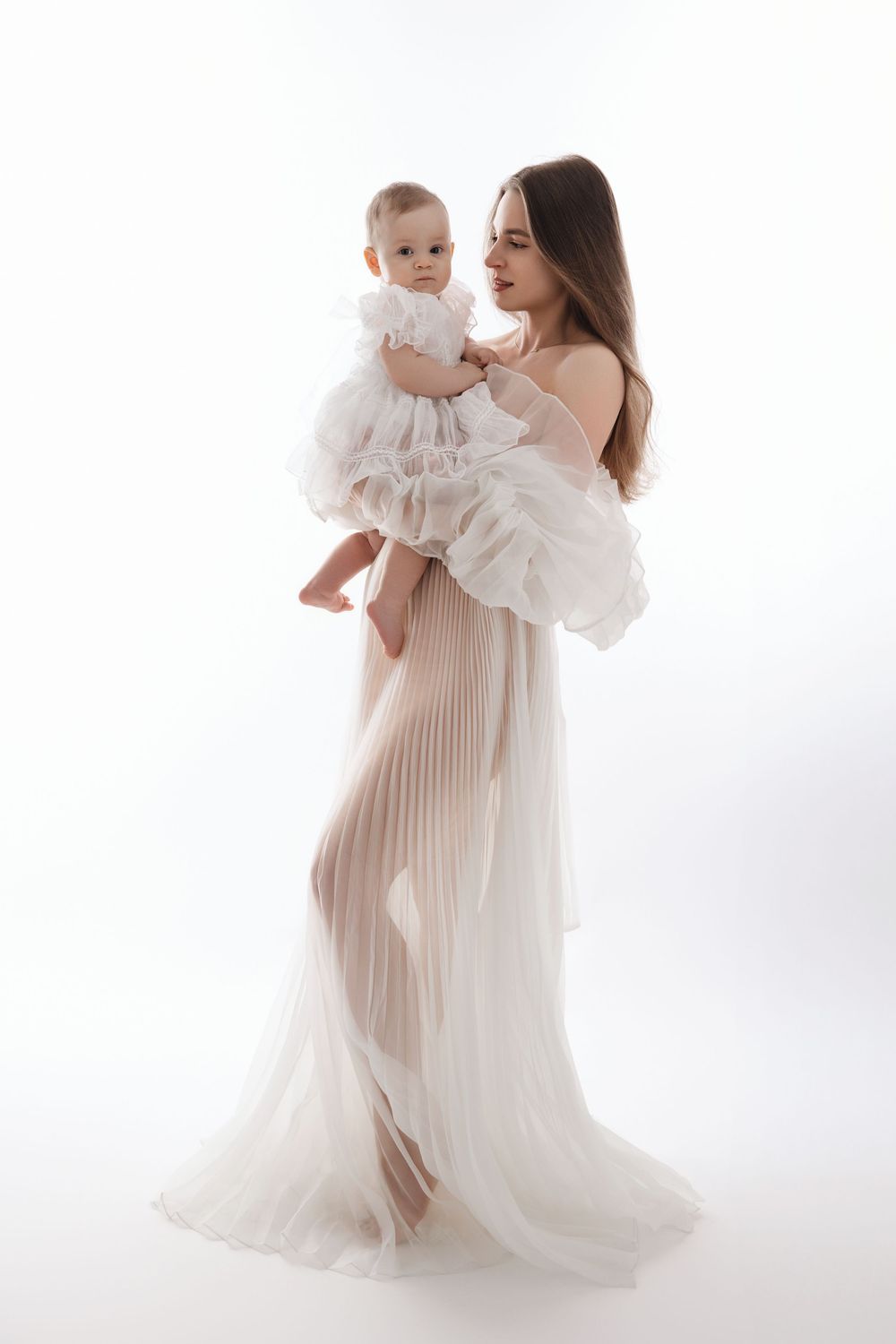 Mother and baby in flowing white dresses embrace against bright studio backdrop in elegant maternity photoshoot.