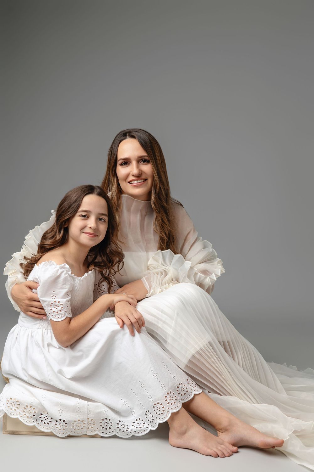 Studio portraits showing tender moments between parent and child in flowing white lace dresses against grey backdrop.