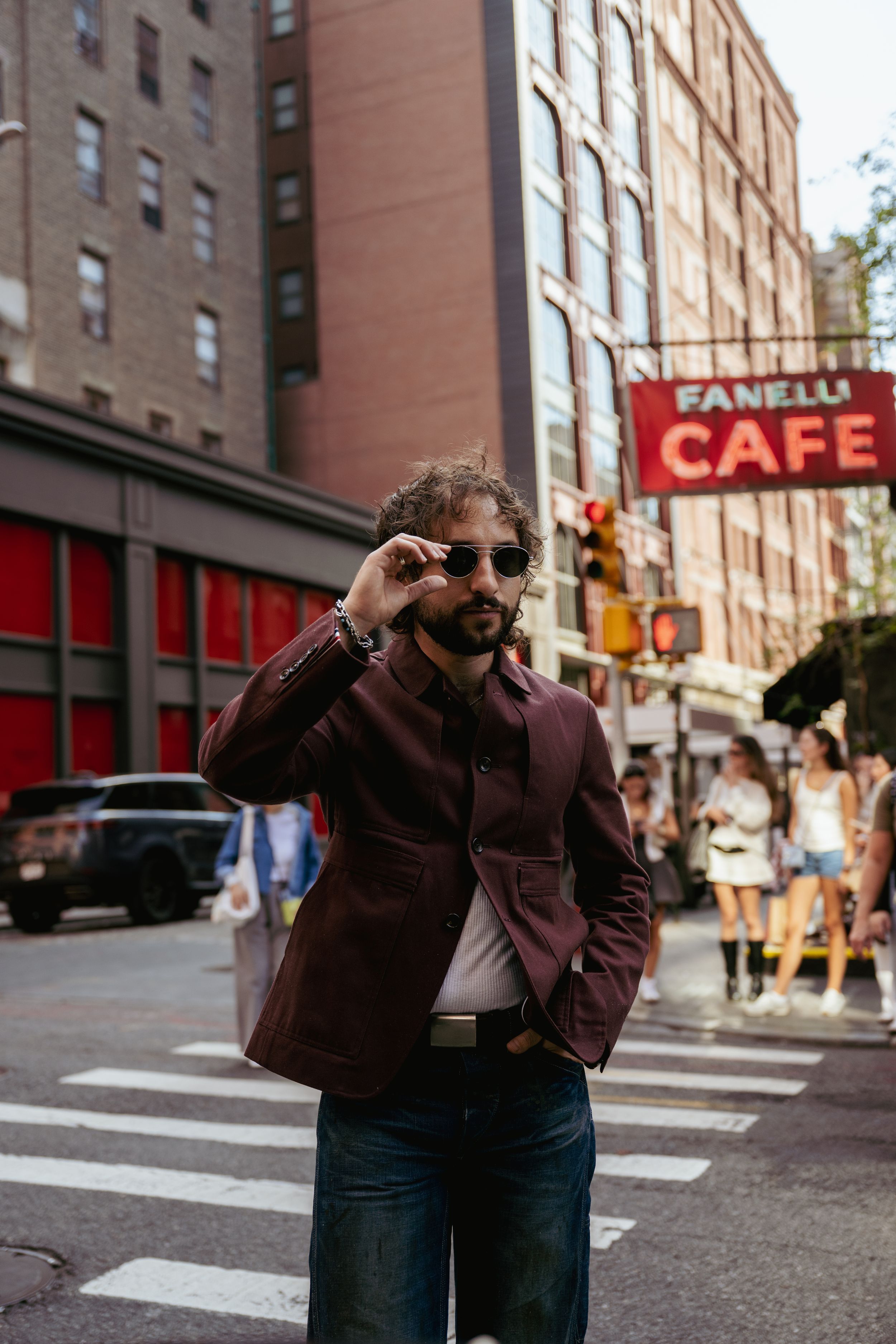 A person in a brown jacket and jeans walks across a city street intersection with brick buildings in the background.
