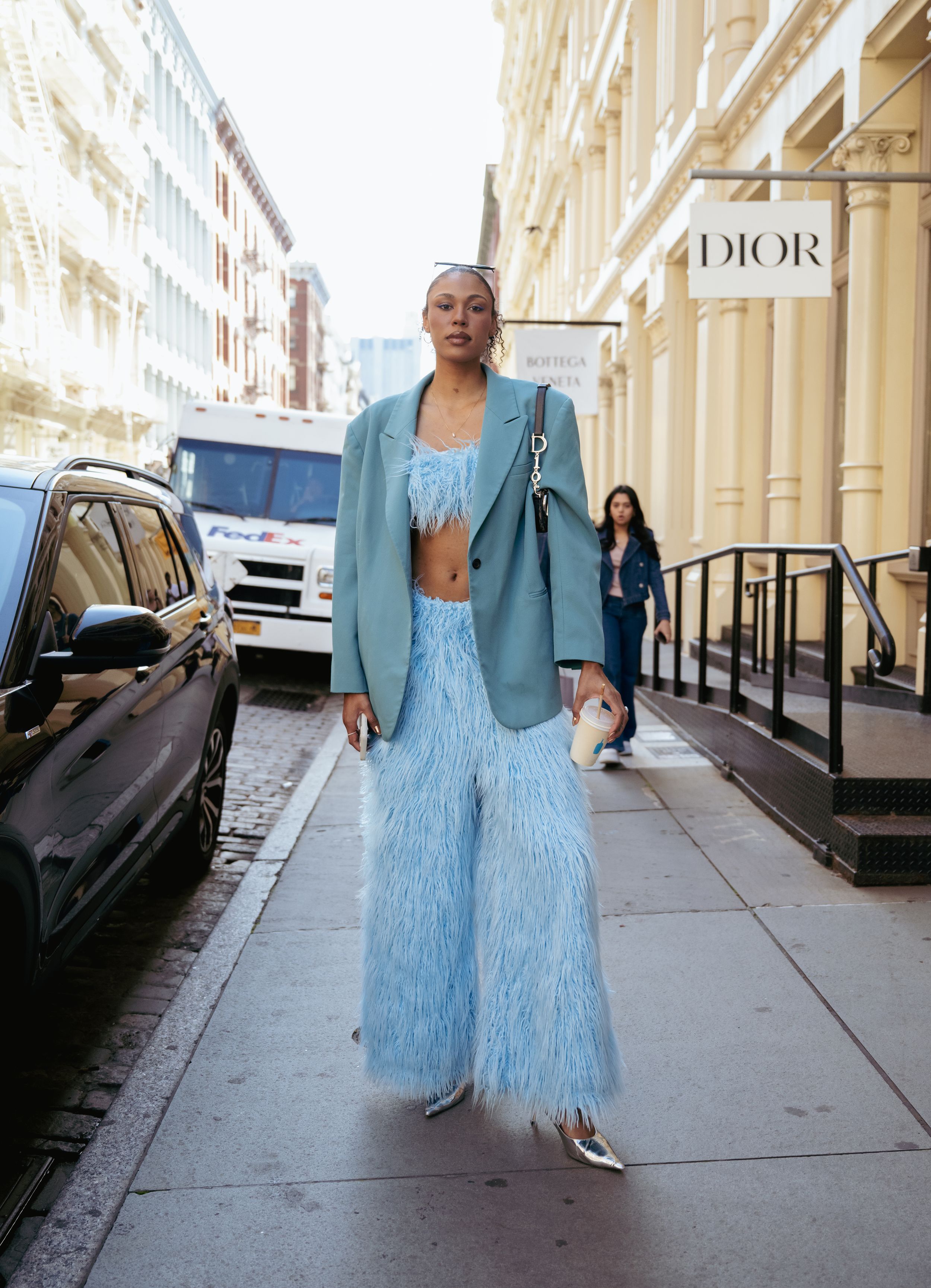 A person wearing a light blue feathered dress and blazer walks down a city sidewalk in multiple sequential shots.