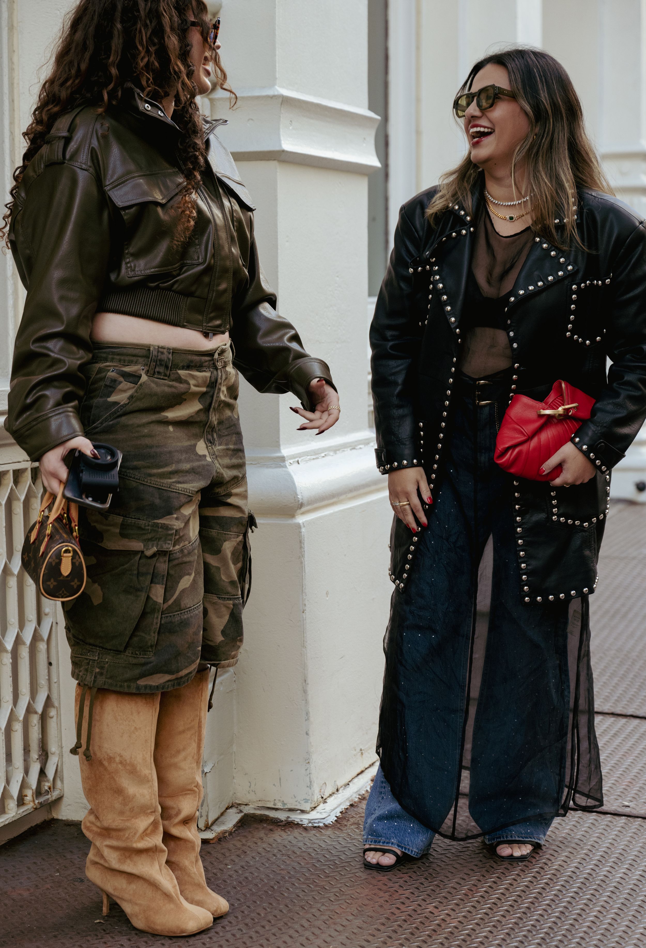 Two people in contrasting outfits chat on the street, one in camo and boots, the other in studded black jacket and wide-leg jeans.