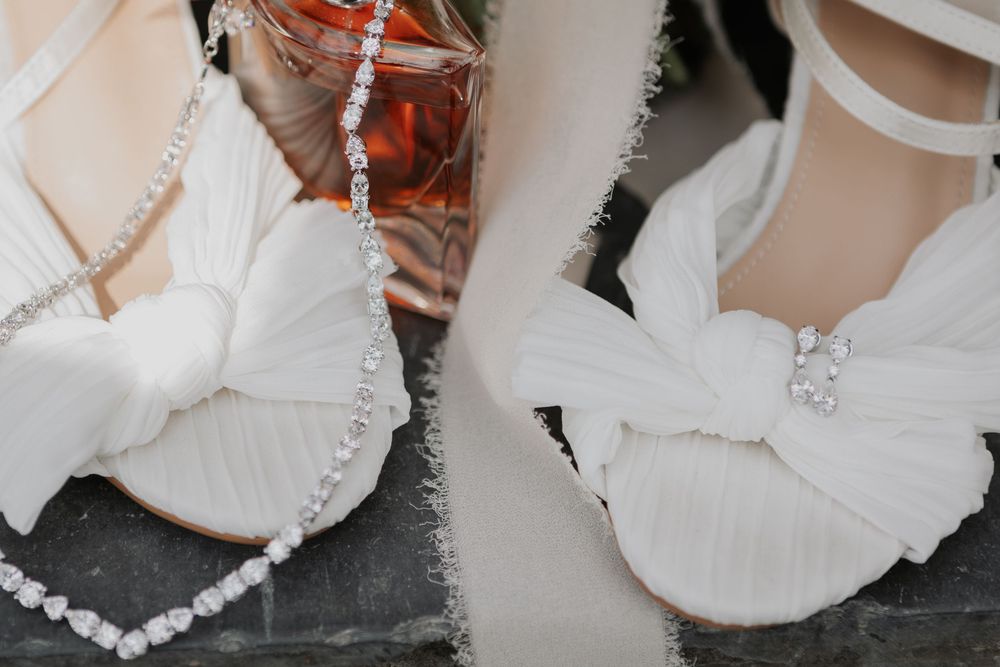 White bridal sandals with bow and crystal embellishment detail displayed alongside a perfume bottle.