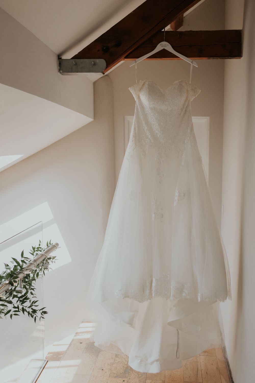 Elegant strapless lace and tulle wedding dress hanging from a wooden beam in a bright airy room.