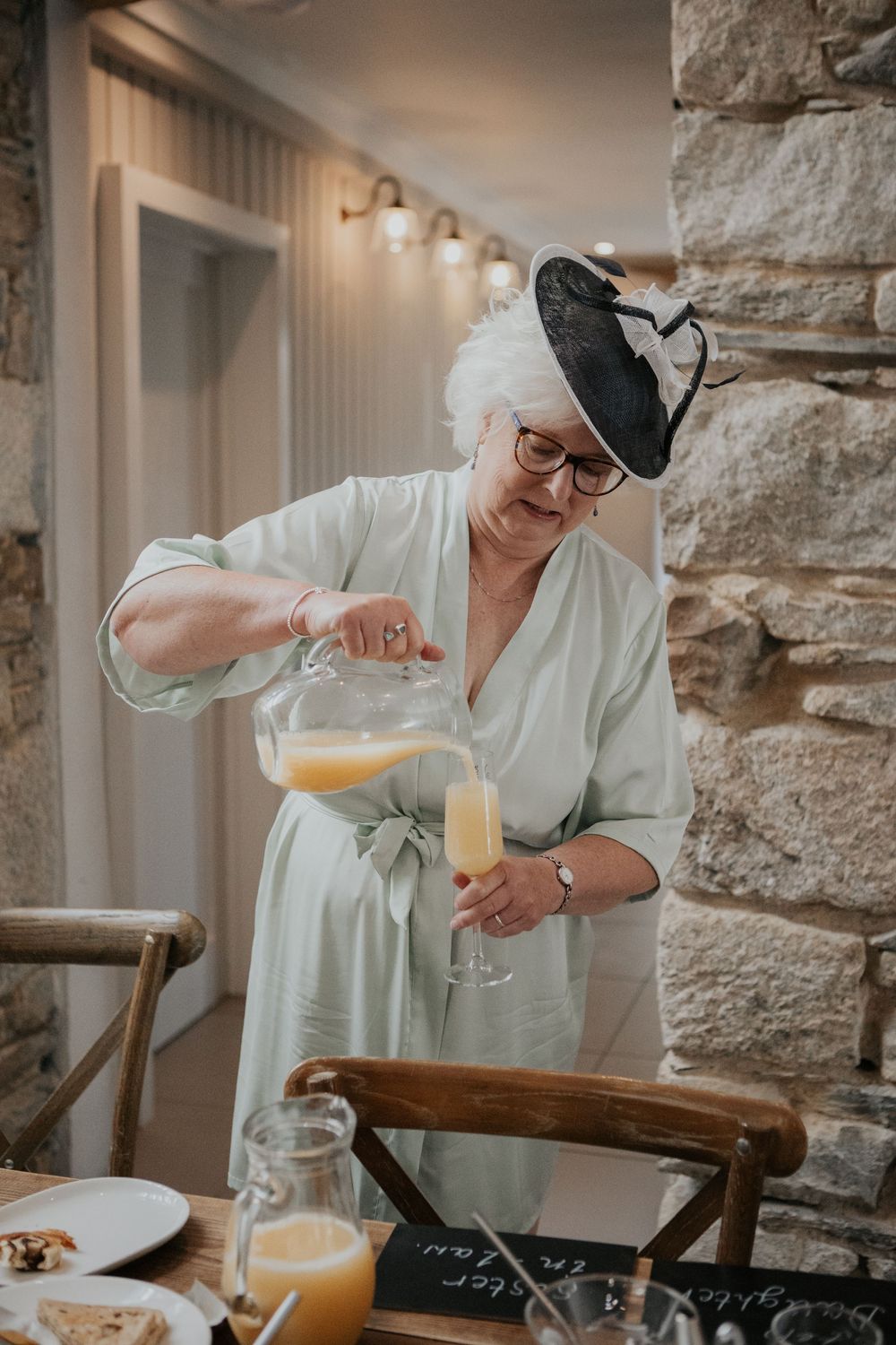 Elegant older woman in sage green robe and fascinator pouring champagne into a flute at a wedding celebration.