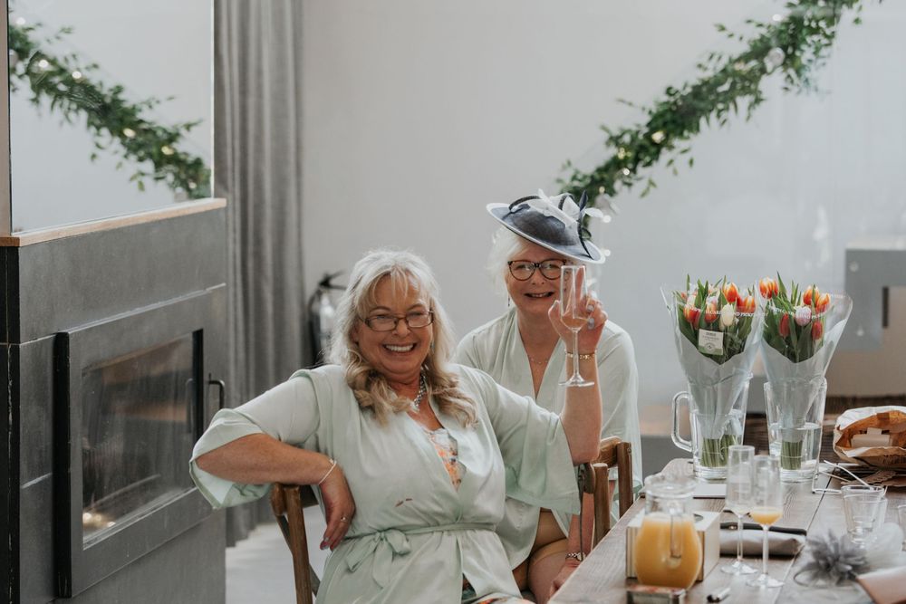 Two smiling older women in matching sage robes and fascinators toasting with champagne glasses at a wedding.