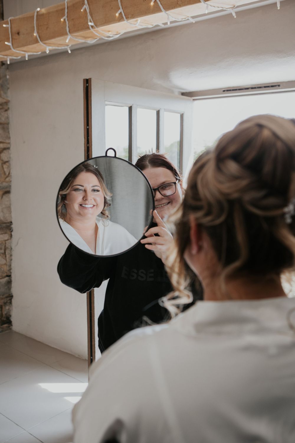 Makeup artist in black robe showing bride her finished look in a round mirror during bridal preparations.