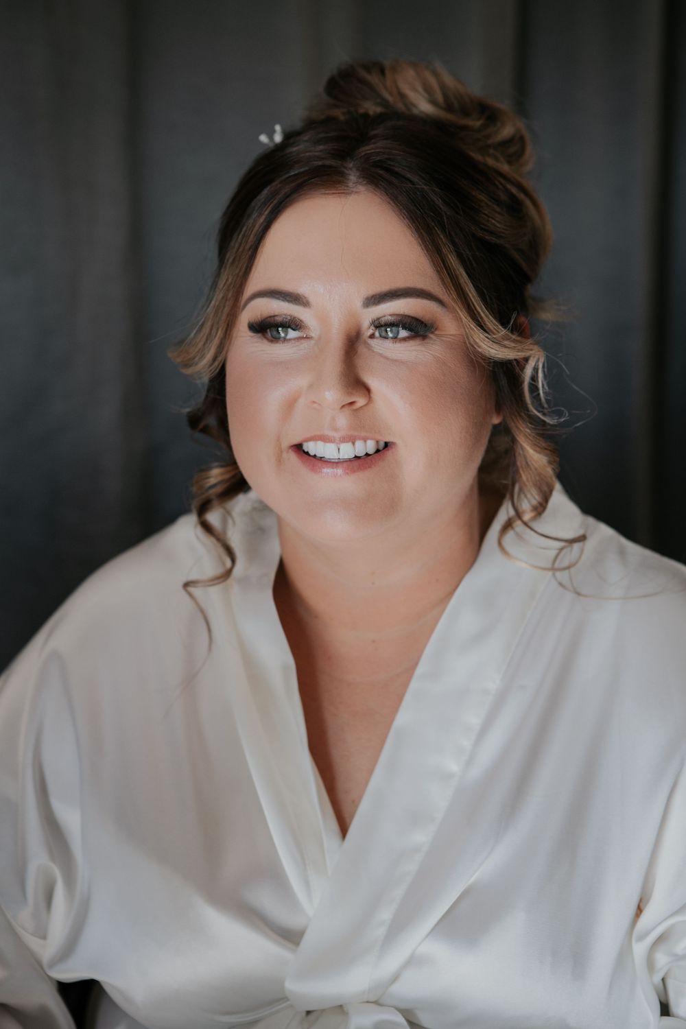 Beautiful bride in white silk robe with elegant updo and natural glam makeup smiling softly.
