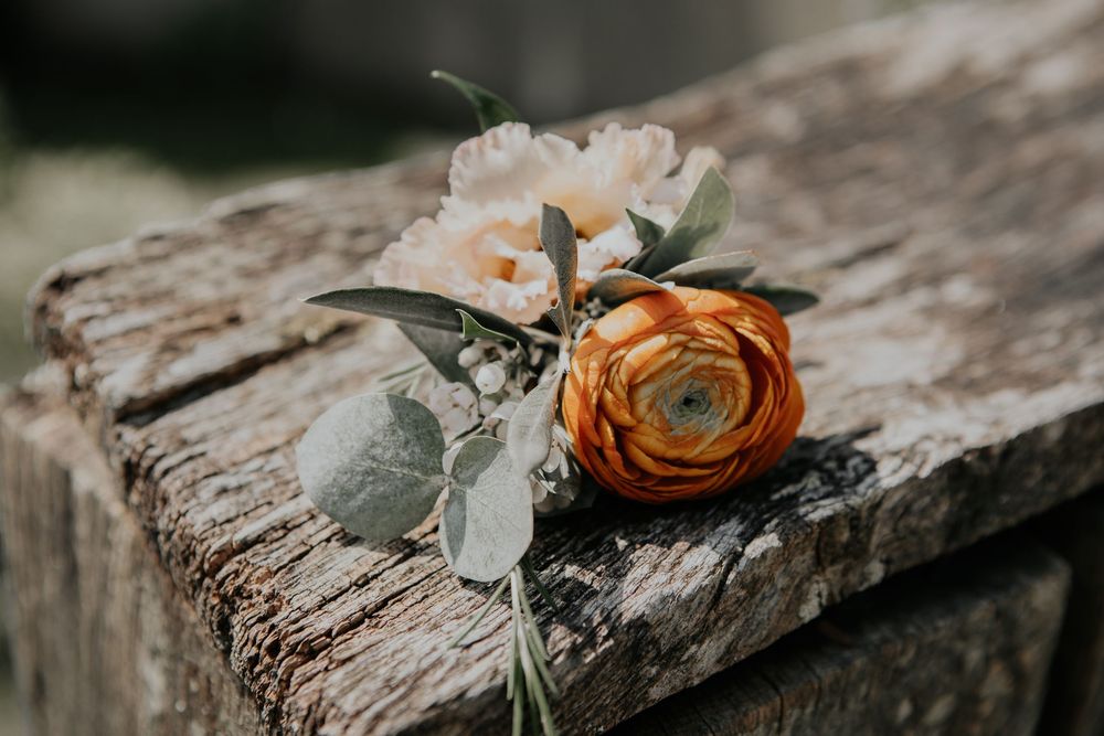 Orange ranunculus and blush flower boutonniere with eucalyptus leaves resting on a rustic wooden surface.