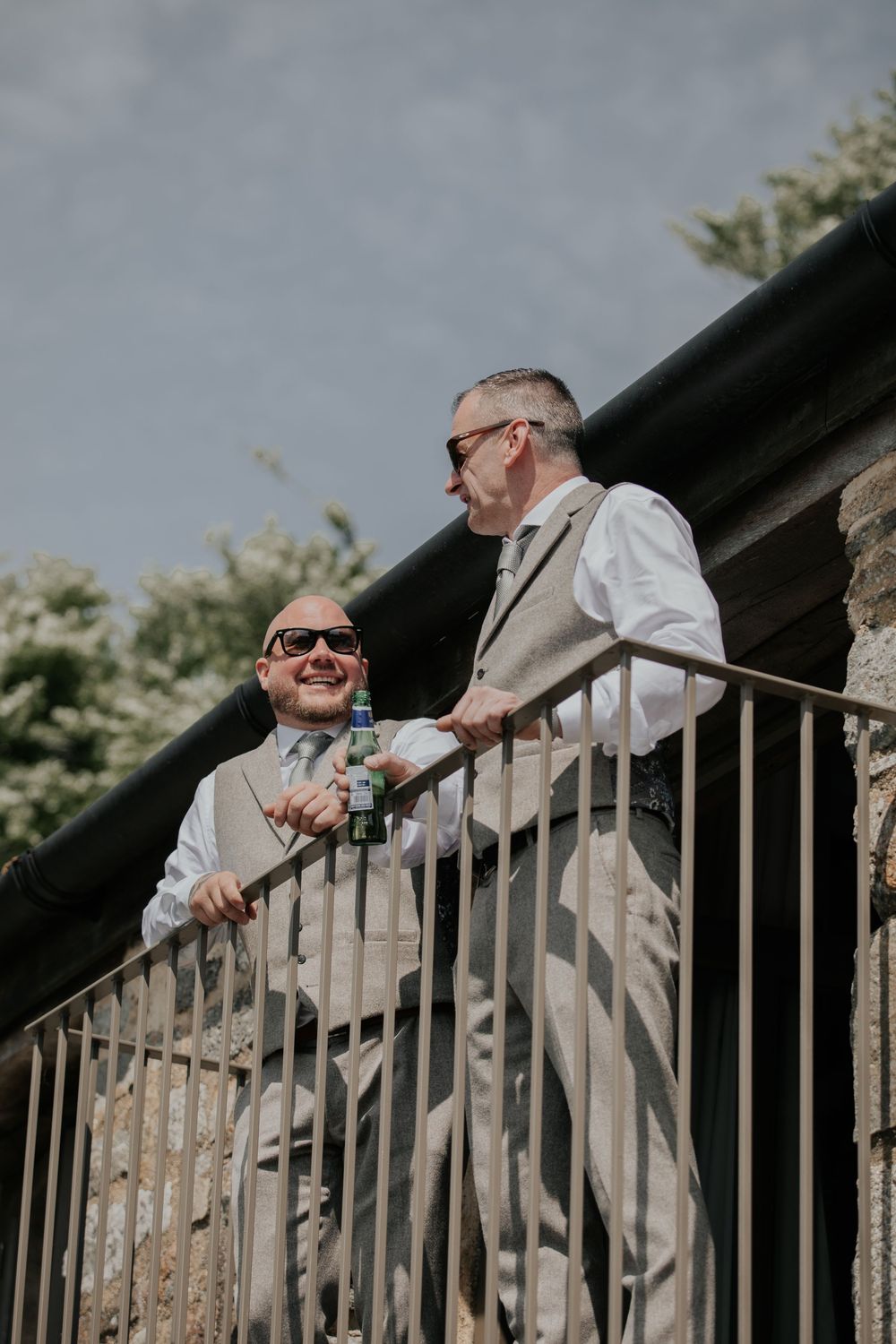 Two groomsmen in grey suits and sunglasses laughing together on a balcony before the wedding ceremony.