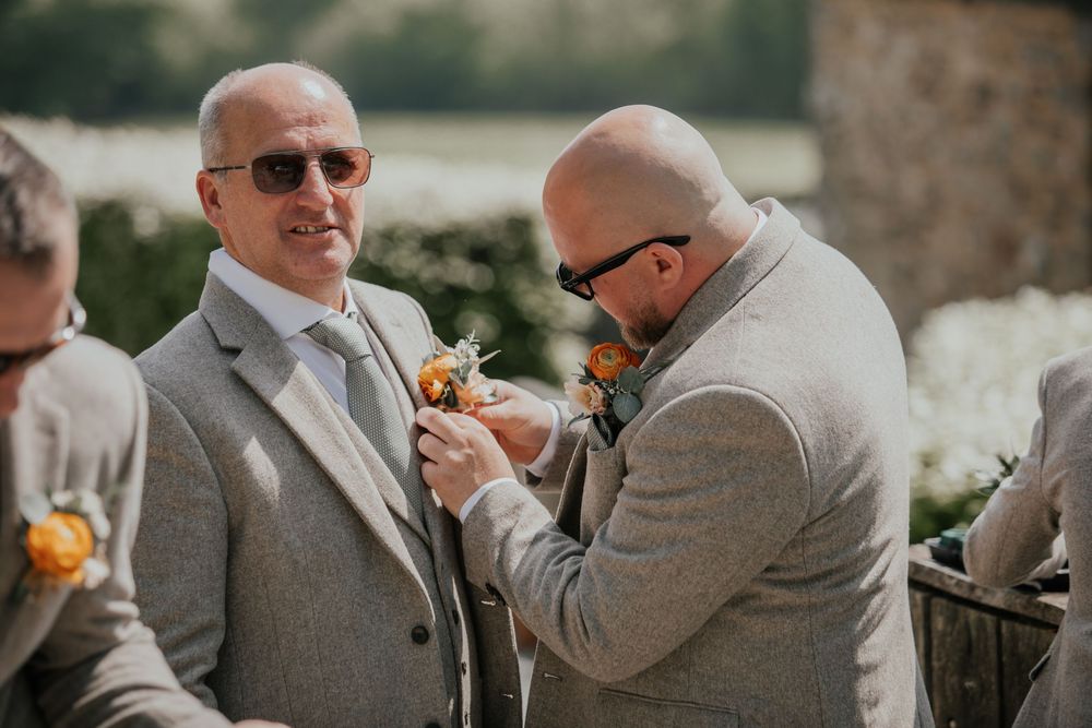 Groomsman pinning an orange floral boutonniere onto the lapel of a smiling groom in a grey suit.