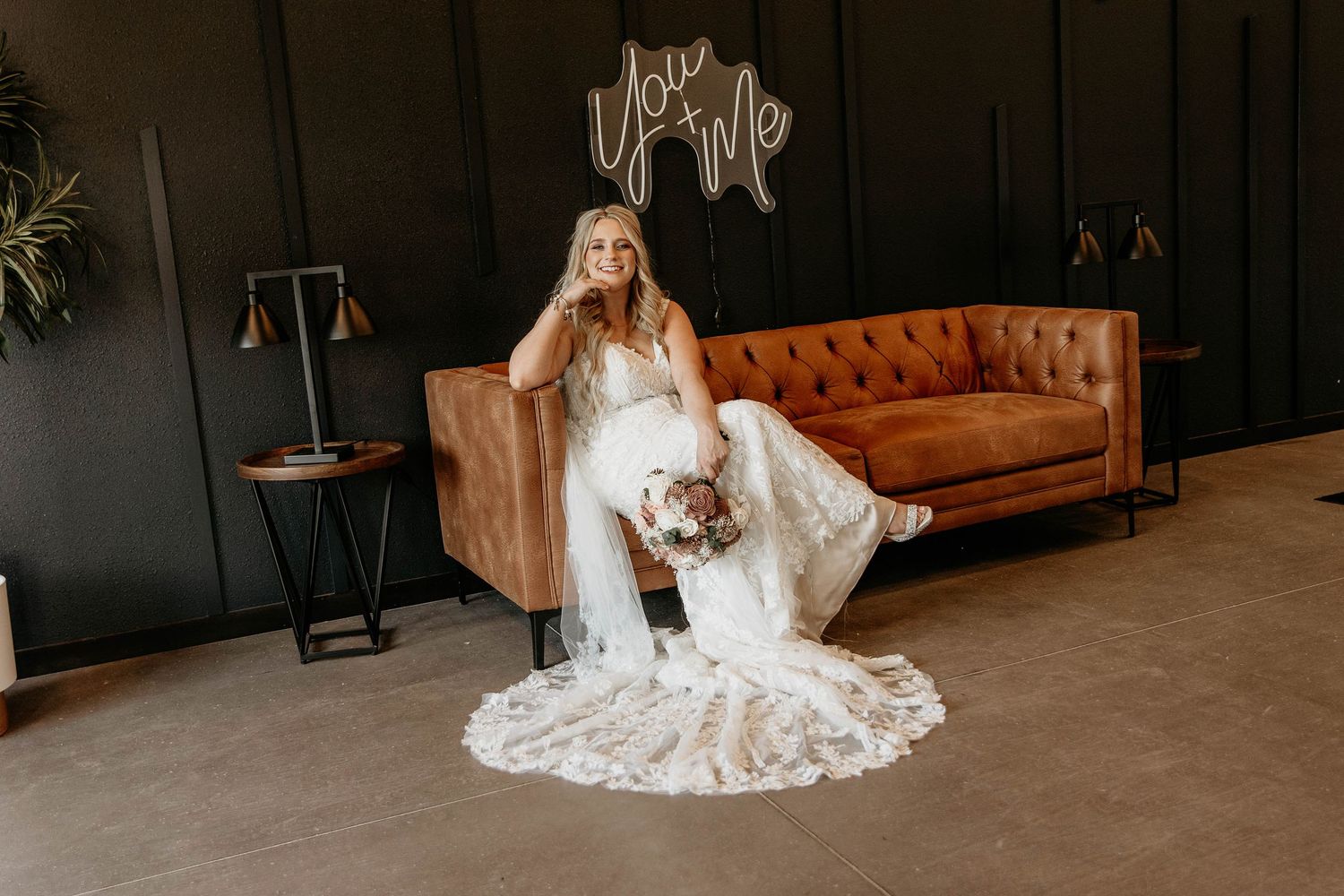 A bride in a flowing white lace wedding dress sits elegantly on a brown leather couch in a dark room.