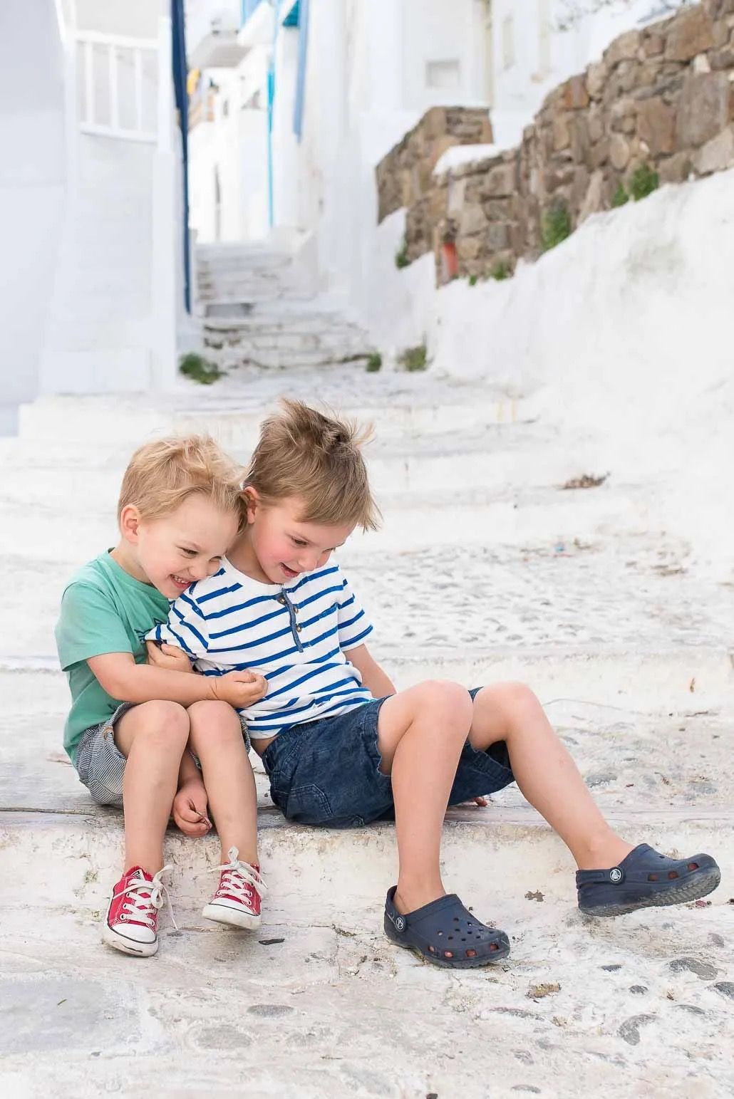 Two young children sitting together on white stone steps in a sunny Mediterranean street.