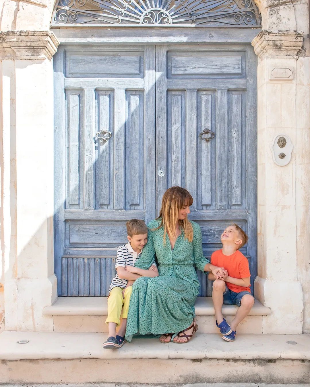 Mother and children sit beside a large blue wooden door on a bright stone street.