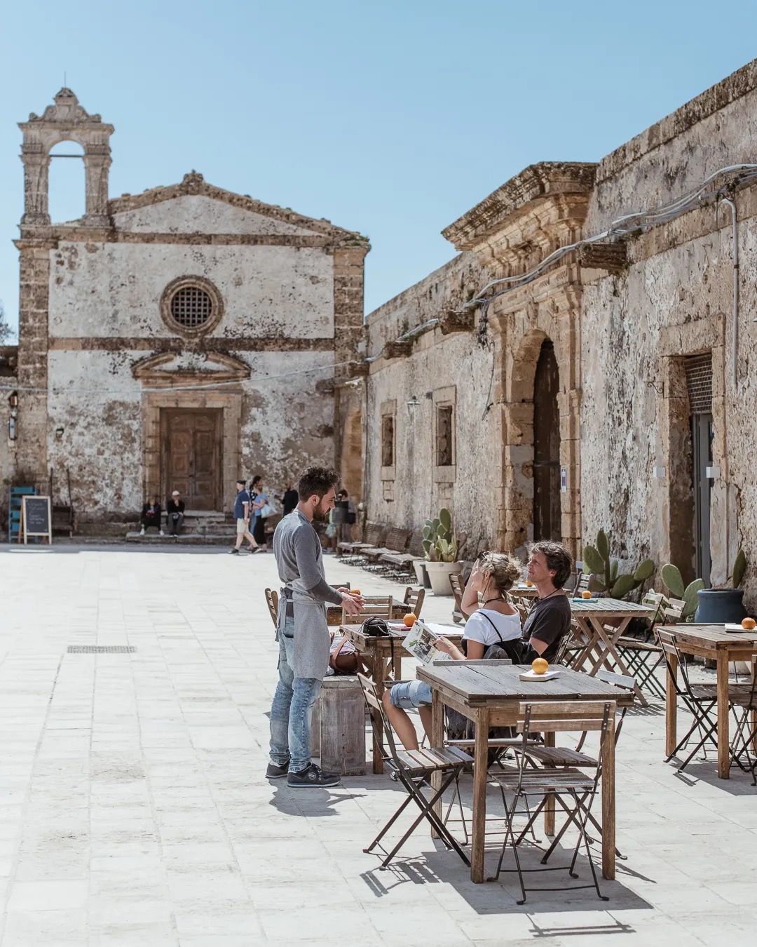 People sit at an outdoor café in a historic stone square beside an old church under bright summer light.