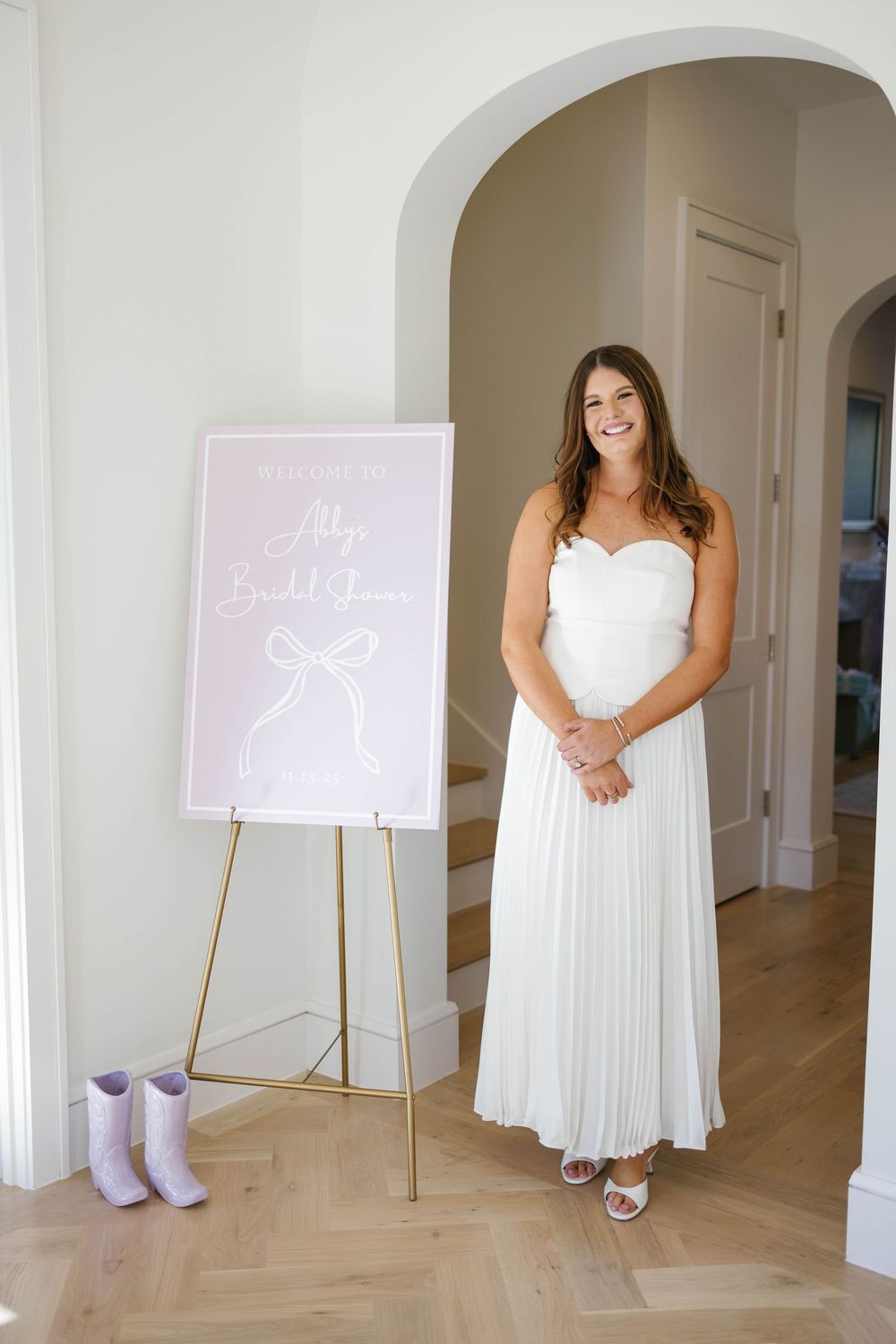 Smiling bride-to-be in white strapless dress standing beside a pink bridal shower welcome sign on a gold easel.