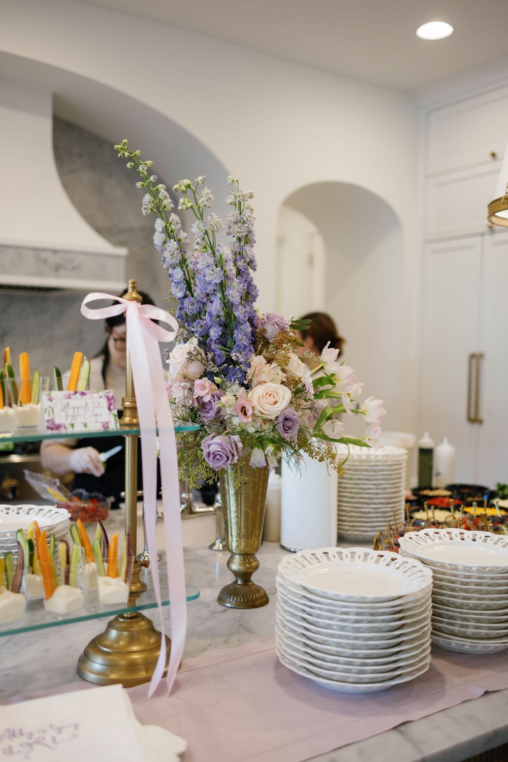 Elegant bridal shower buffet with purple and blush floral arrangement, stacked plates, and desserts on a marble counter.